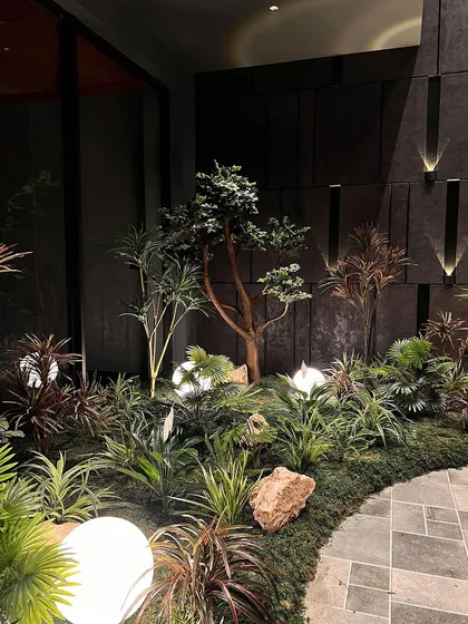 A wide view of an indoor garden featuring a variety of artificial plants, from small bonsai to taller, leafy specimens. The glowing globe lights and mossy ground cover create a complete, immersive landscape.