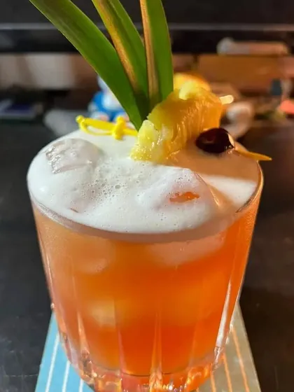 A tropical orange-colored cocktail with a frothy top, garnished with pineapple and a cherry. Perfect for a summer event or a beach-themed party.
