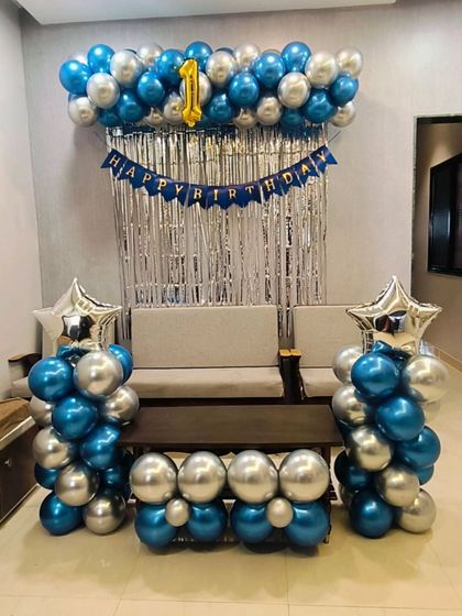 A classic birthday decoration for a home celebration, featuring blue and silver balloon pillars, a shimmer curtain, and a "Happy Birthday" banner.