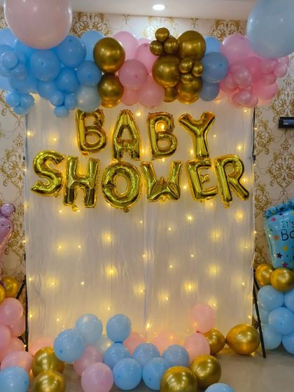 A simple and sweet baby shower backdrop. This design uses a white curtain with fairy lights, 'BABY SHOWER' foil balloons, and pink and blue balloon pillars, perfect for a gender reveal theme.