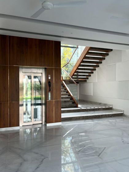 The foyer at Gulmohar Farmhouse features a glass elevator clad in wood paneling, situated next to the modern, open-riser staircase. The design is a study in warm minimalism.