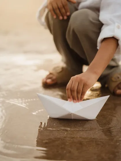 A close-up of a boy's hand placing a paper boat in the water. This detailed shot tells a story of childhood innocence and play.
