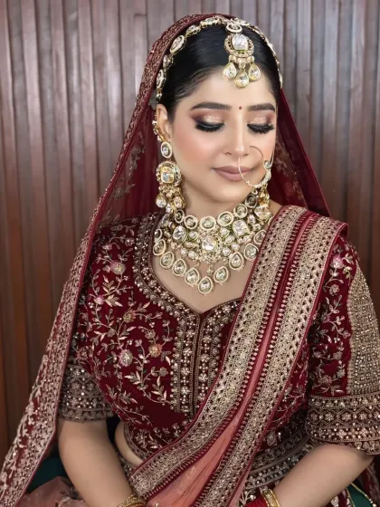 A bride in a deep maroon velvet lehenga. The makeup is soft and elegant, with a focus on a flawless base and a subtle nude lip to balance the heavy outfit.