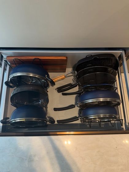 A drawer organized with stacks of non-stick pans and lids, using a rack to keep them upright and prevent scratching. The wooden chopping board has its own dedicated space.