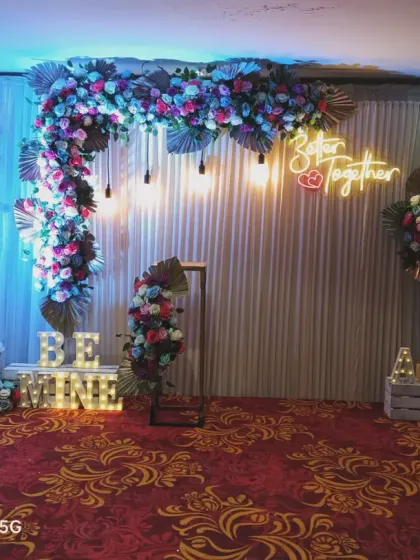 A similar design adapted for a larger venue. This engagement backdrop features a floral arch with dried palm leaves, "Better Together" and "Be Mine" signs, and is set on a patterned carpet.