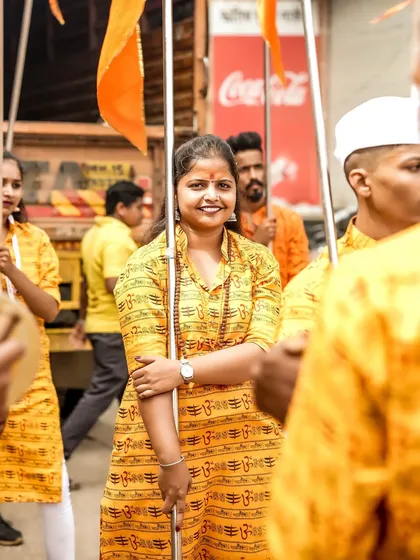 A smiling member of the dhol tasha troupe, holding a flag and posing for a photo amidst the procession.