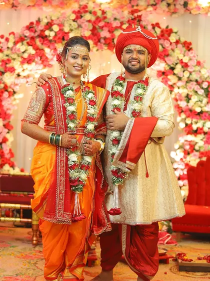 A classic portrait of the happy couple against a floral backdrop. The bride's orange and red Nauvari saree is a traditional and vibrant choice for her wedding day.