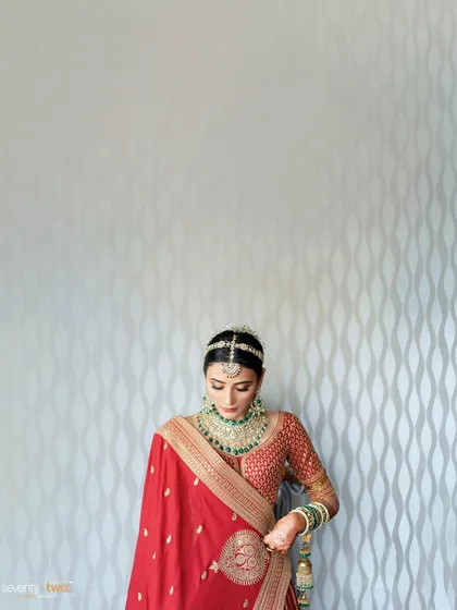 A bride in a red saree stands against a textured white wall, a simple yet elegant composition that highlights the richness of her attire.