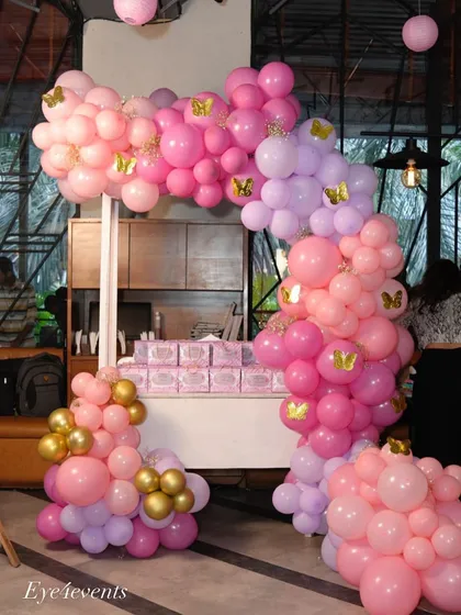 A whimsical candy cart decorated for a baby shower or birthday party. The cart is framed by a large balloon arch in shades of pink, purple, and gold, with small golden butterfly details adding a magical touch.