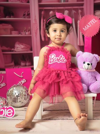 This toddler, dressed in a pink Barbie outfit, sits confidently with her purple teddy bear, creating a sweet and stylish portrait.