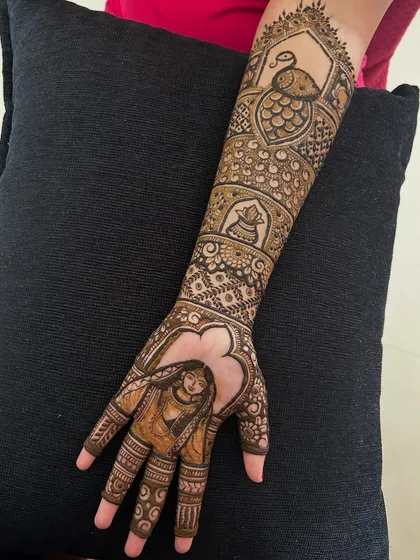 A full-arm design that incorporates a bride portrait, a peacock, and a traditional pot (kalash) motif.