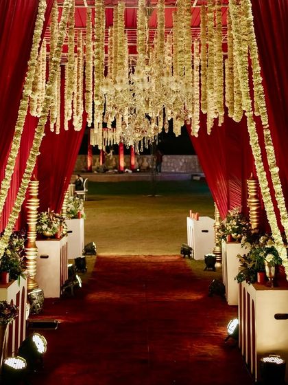 A grand entrance for a red-themed reception, with a walkway draped in rich red fabric and lined with hanging strings of white flowers, leading into the main event space.