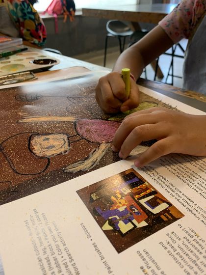 A student uses a yellow crayon to fill in parts of their drawing, with reference materials visible nearby.