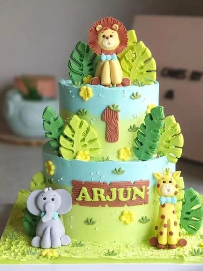 A two-tier jungle theme cake with cute fondant animals. The design features a lion, elephant, and giraffe, surrounded by tropical leaves.