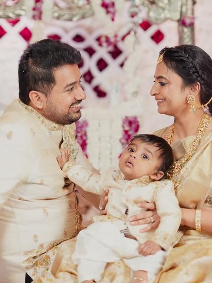 A tender family moment. The parents and their baby are dressed in coordinated cream and gold outfits, with delicate embroidery on the father's and baby's kurtas matching the mother's elegant silk saree.