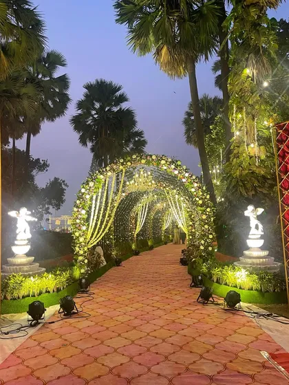 A magical garden entrance at dusk. The floral archway is adorned with cascading fairy lights, and the path is flanked by glowing cupid statues, creating a truly romantic and enchanting atmosphere.