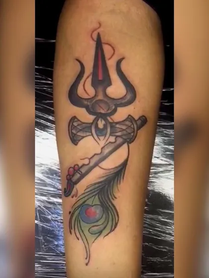 A creative fusion of spiritual symbols. This tattoo combines Lord Shiva's Trishul with Lord Krishna's flute and peacock feather. The use of color highlights the different elements of this unique divine concept.