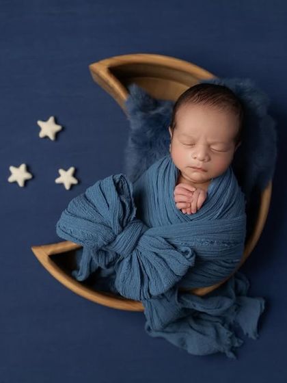 A newborn boy wrapped in a deep blue swaddle, resting in the crescent of a wooden moon prop against a matching blue background. The tiny stars complete this celestial theme.