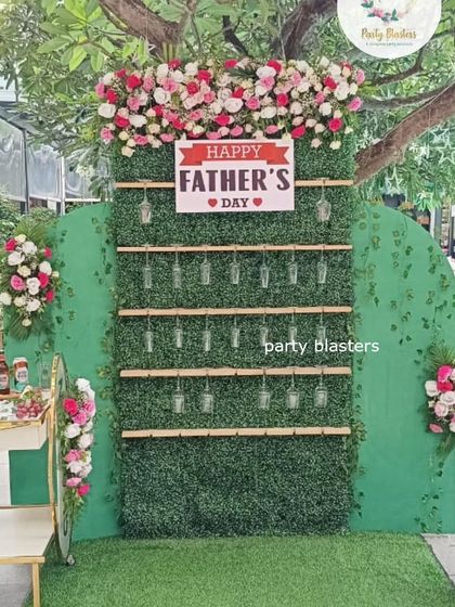 A creative "Cheers to Dad" wall for a Father's Day celebration. The green hedge backdrop is set up to hold drink glasses for guests, topped with a floral arrangement and a "Happy Father's Day" sign.