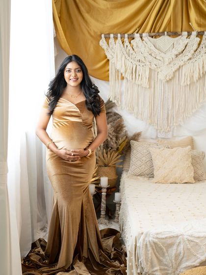 A full-length portrait of an expecting mother in a golden velvet gown. She stands in a beautifully decorated boho room, smiling warmly at the camera.