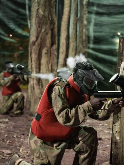 A paintball player kneels behind cover, smoke billowing from his marker as he takes a shot. The action is real.