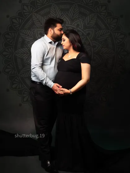 A loving couple's portrait set against a dark, patterned backdrop. The father-to-be kisses his partner's head as they both gently hold the baby bump, creating a deeply emotional image.