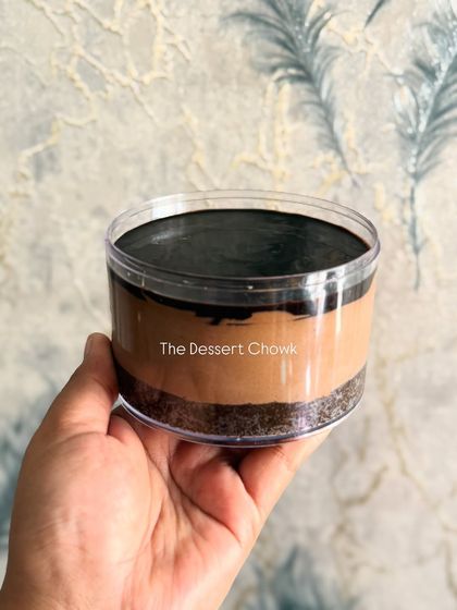 A side view of the no-bake chocolate cheesecake tub, showing the distinct layers of biscuit crust, creamy chocolate cheesecake filling, and a dark chocolate ganache on top.