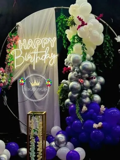 A beautiful birthday decoration with a circular frame, a balloon garland in purple, silver, and white, and a 'Happy Birthday' neon sign.
