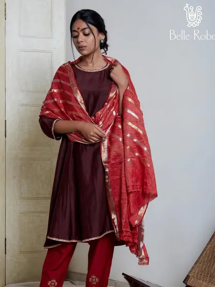 A classic look featuring a deep maroon kurta set with a contrasting red dupatta. The makeup is traditional and elegant, with a defined eye and a red bindi, perfect for a puja or a family gathering during wedding festivities.