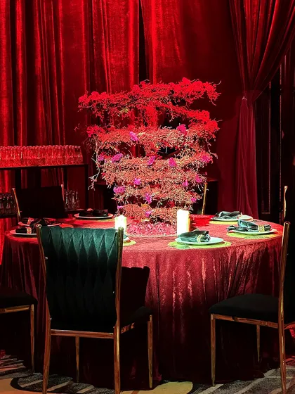 An intimate dining table setup, draped in red velvet and featuring a sculptural red floral centerpiece, surrounded by deep green chairs for a rich, jewel-toned look.