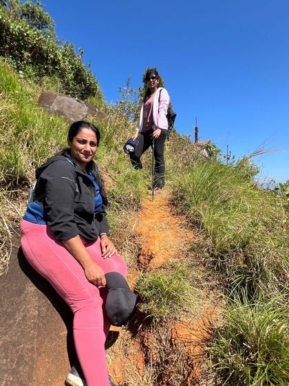 Two trekkers on the trail to Kurinjal Peak, with the path winding through the green hills.
