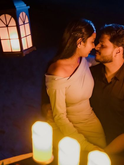 An intimate moment from a romantic dinner shoot. The couple leans in close, illuminated by the soft glow of candles and a lantern, creating a warm and passionate atmosphere.