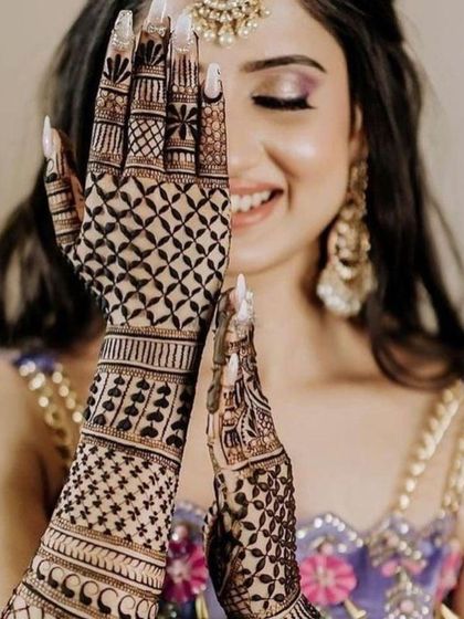 A joyful bride peeking through her intricately designed hands. The design features a mix of geometric jaali work and delicate heart-shaped patterns on the arms.