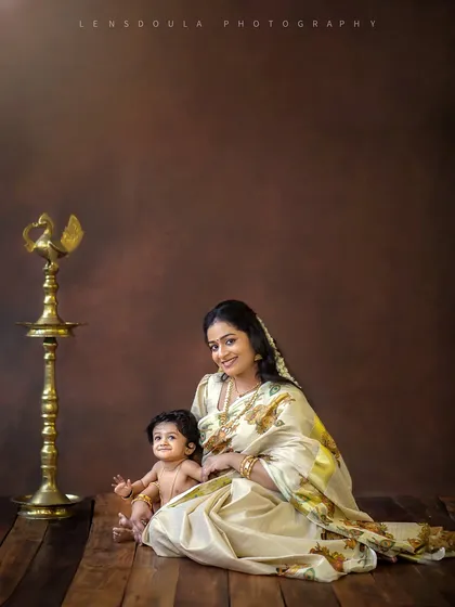 A stunning portrait of a mother and her baby in a traditional 'Mallu' themed setup. The rich brown backdrop and the classic brass lamp (Nilavilakku) add to the authenticity and warmth.