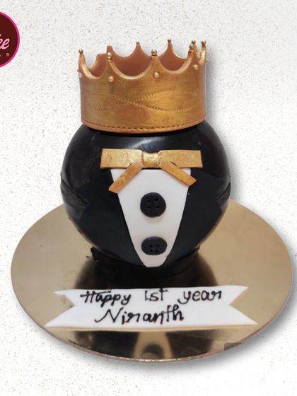 A stylish tuxedo-themed smash cake for a first birthday. The round cake is decorated to look like a little suit with a crown on top, perfect for a "little prince" theme.