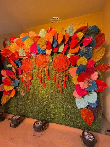 A fun and creative photo backdrop for a Mehendi, made with a green hedge wall, colorful paper fans, and bright red dreamcatchers.