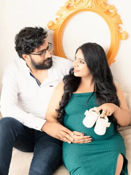 A cozy and sweet moment. The couple sits together, holding a pair of tiny baby shoes, their expressions full of warmth and anticipation.