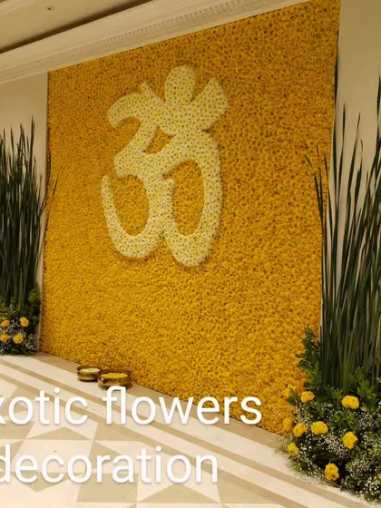 A serene and powerful statement wall featuring a large Om symbol crafted from white chrysanthemums set against a backdrop of vibrant yellow marigolds. Flanked by tall green foliage, this floral installation creates a peaceful and spiritual ambiance for meditation spaces and religious events.
