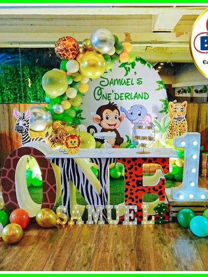 A "Samuel's Onederland" jungle party setup. This decor features a large 'ONE' table with animal prints, character cutouts, and a green and gold balloon arch.