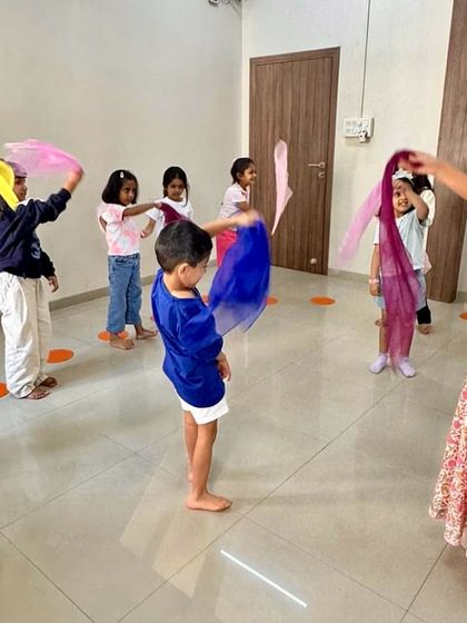 A teacher leads a group of children in a scarf dance. These activities help develop gross motor skills, coordination, and the ability to follow musical cues.
