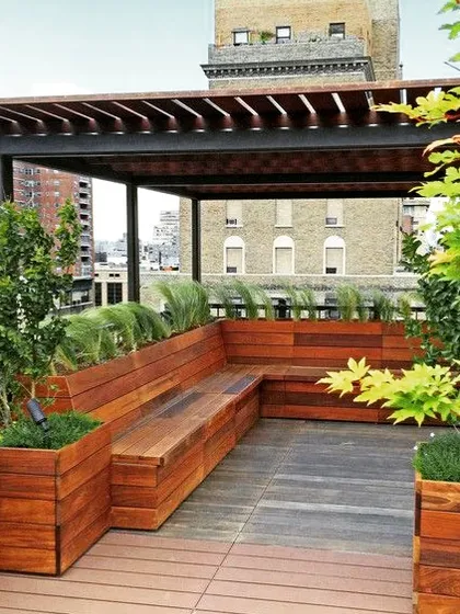 This urban rooftop deck features a pergola with built-in wooden planter boxes that double as bench seating. The design maximizes a small footprint by integrating greenery and seating into a single cohesive structure.