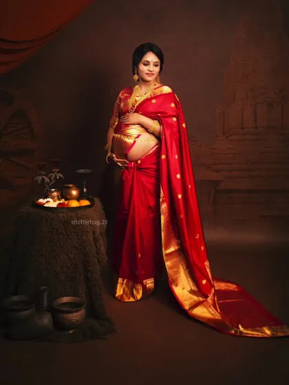 Another angle from our traditional saree shoot, showing the full setup with antique-style props and a temple-themed backdrop. The mom-to-be looks radiant holding her bump.