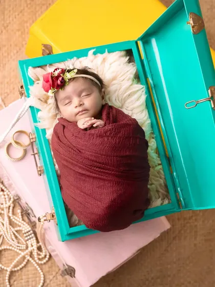 A close-up of a newborn sleeping in a colorful trunk. The pearls and flowers add a touch of vintage glamour to the scene.