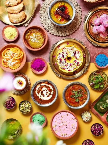 A colorful Holi feast spread, featuring a variety of traditional Indian dishes and sweets. The scattered colored powders and floral elements perfectly capture the vibrant spirit of the festival.