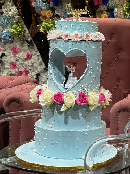 This breathtaking three-tier engagement cake features a unique heart-shaped cutout, revealing a delicate figurine of the couple. The soft blue frosting, pearl details, and fresh roses create a truly romantic and unforgettable centerpiece for a special engagement party.