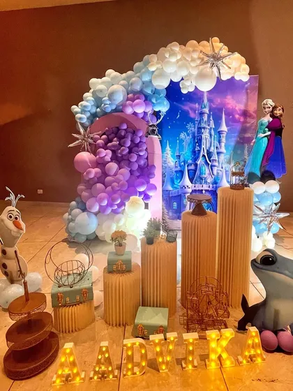 A magical Disney 'Frozen' party setup for Aadvika. This wide shot shows the stunning Arendelle castle backdrop, an elaborate balloon arch in shades of purple, blue, and white, and character cutouts that bring the winter wonderland to life.