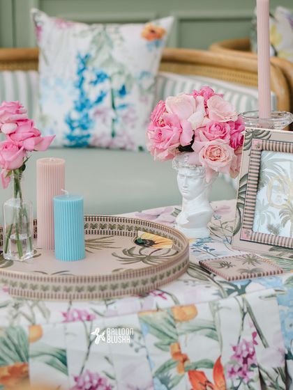 A close-up on a tabletop, showing how we layer different elements. A custom-printed tray holds pink roses in a classic bust vase, flanked by colorful candles and a patterned picture frame, all set against floral linens.