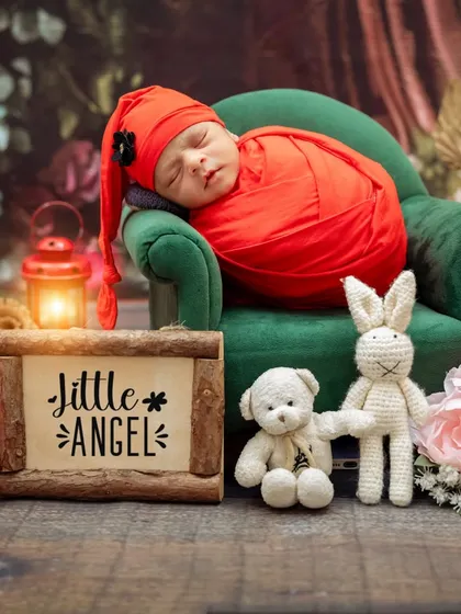 A newborn sleeps on a miniature sofa in a red-themed setup, complete with a "Little Angel" sign and other cozy props.