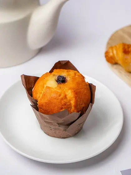 A blueberry muffin in its paper case, styled with a croissant and a teapot in the background. This creates a complete breakfast or tea-time scene.