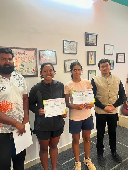 Teammates Paavani Bisht and Eshanya Khattar celebrate together after their AITA CS National U-16 final. This indoor photo shows them with their coaches, holding their winner and finalist certificates.